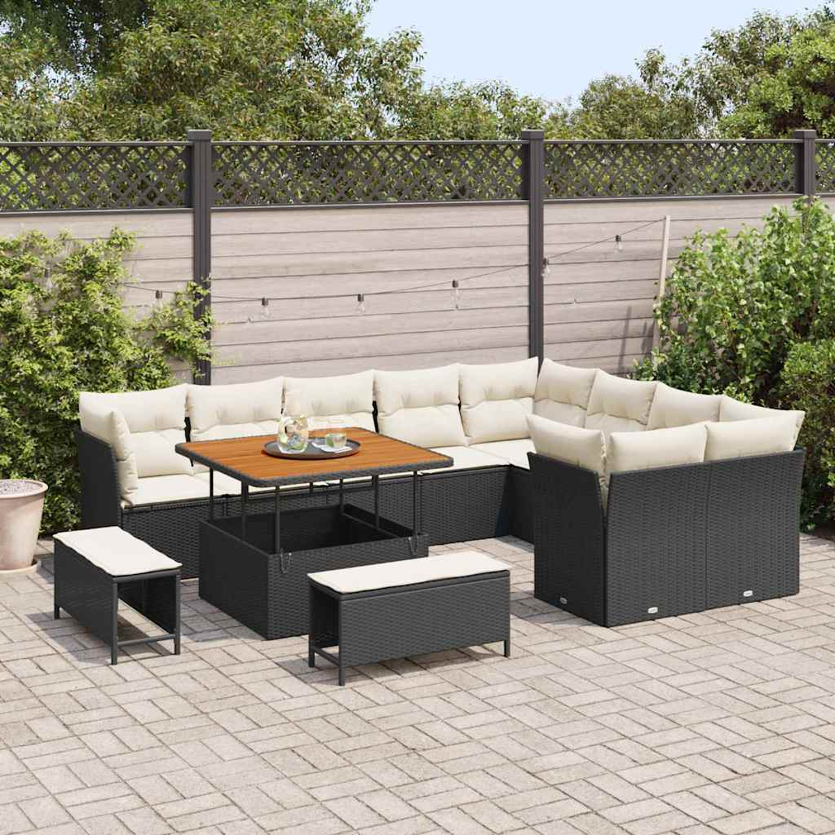 Garten-Set mit schwarzer Korbmöbeln, beigen Kissen, Holztisch und Steinterrasse. Im Hintergrund ein Zaun und Grün.