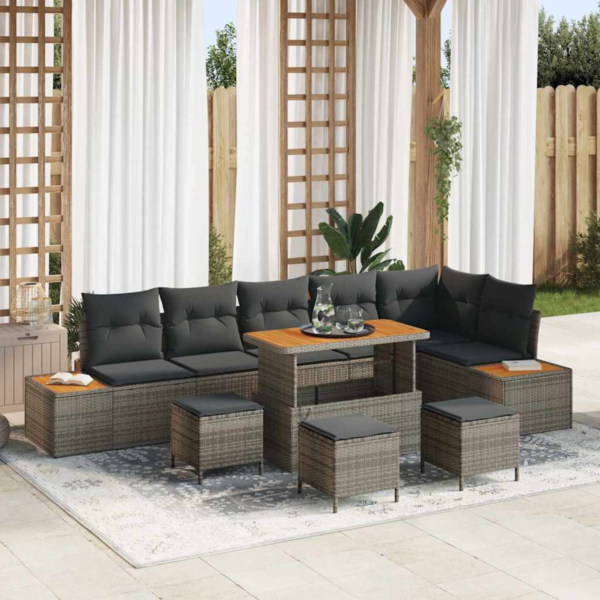 Set da patio: divano in vimini grigio con cuscini neri, tavolo e sgabelli in legno, su un tappeto, tende bianche e recinzione in legno.