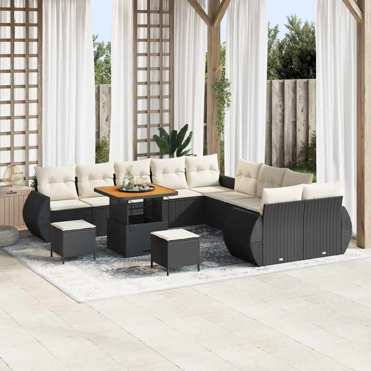 Zona de estar al aire libre con sofá de ratán negro, cojines beige, mesa de madera y taburetes. Cortinas blancas y estructura de madera.