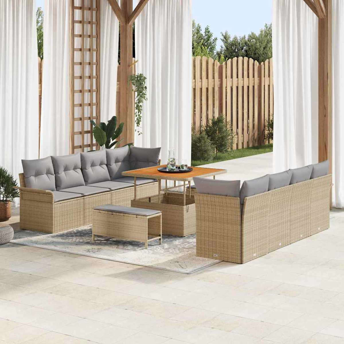 Set da patio all'aperto con mobili in vimini beige, cuscini grigi, tavolo in legno e tende bianche. Recinzione in legno e vegetazione sullo sfondo.