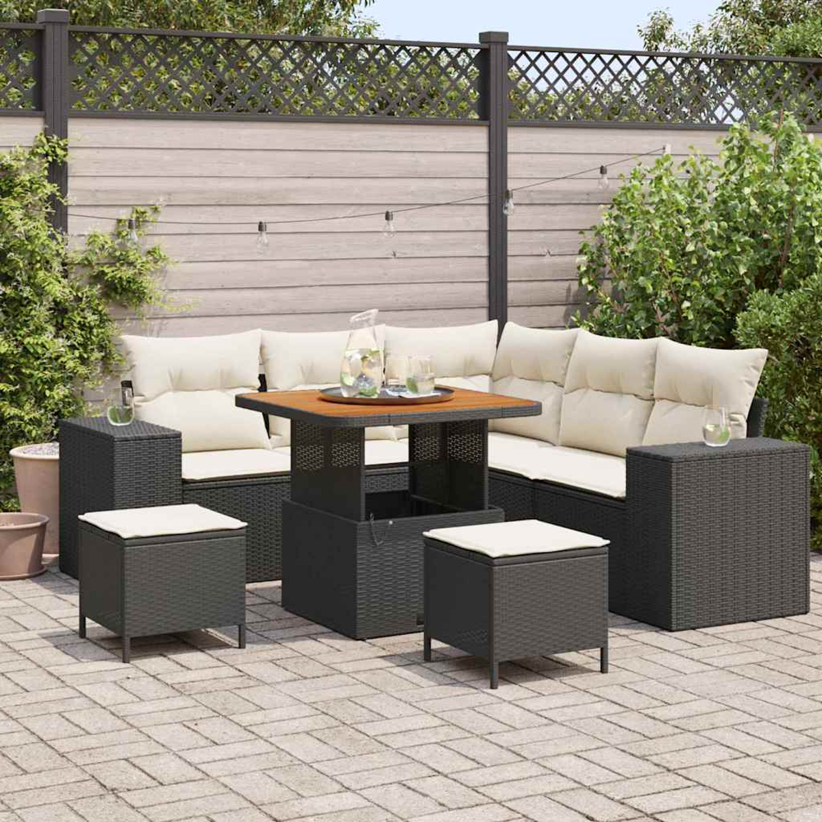 Set da patio: divano angolare con cuscini color crema, tavolo in legno, due pouf e due tavolini. Patio in mattoni con recinzione in legno.
