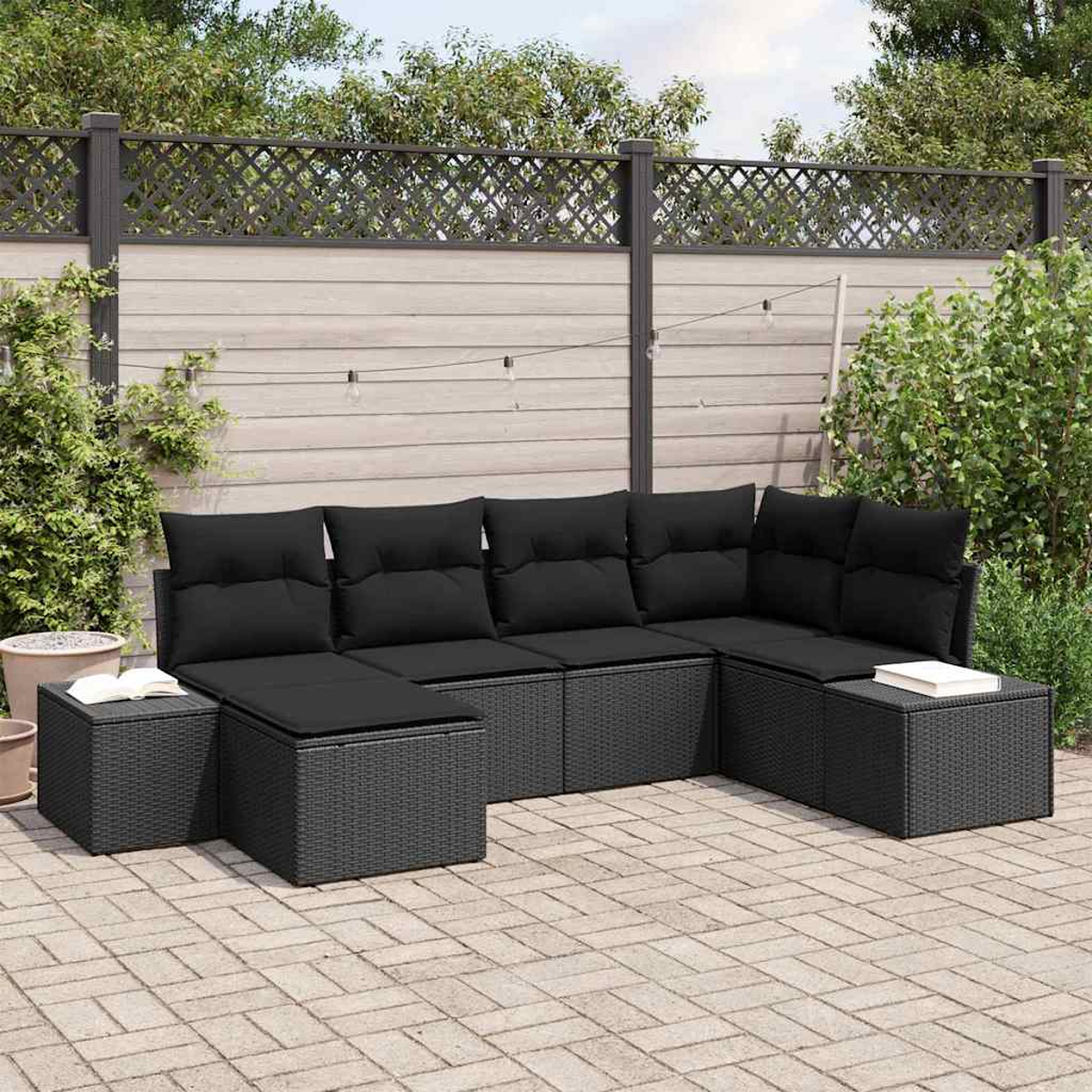 Divano angolare in rattan nero con cuscini su un patio in mattoni. Lo sfondo comprende una recinzione, piante e luci a stringa.
