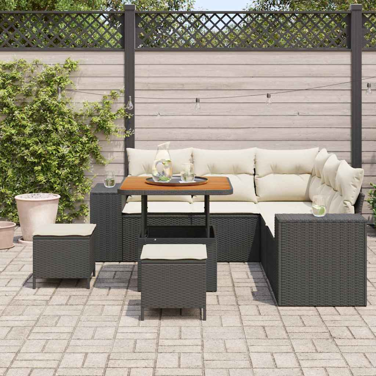 Set da patio esterno: divano, tavolo e sgabelli in vimini grigio scuro con cuscini color crema, su un patio in mattoni. Sfondo: recinzione in legno.