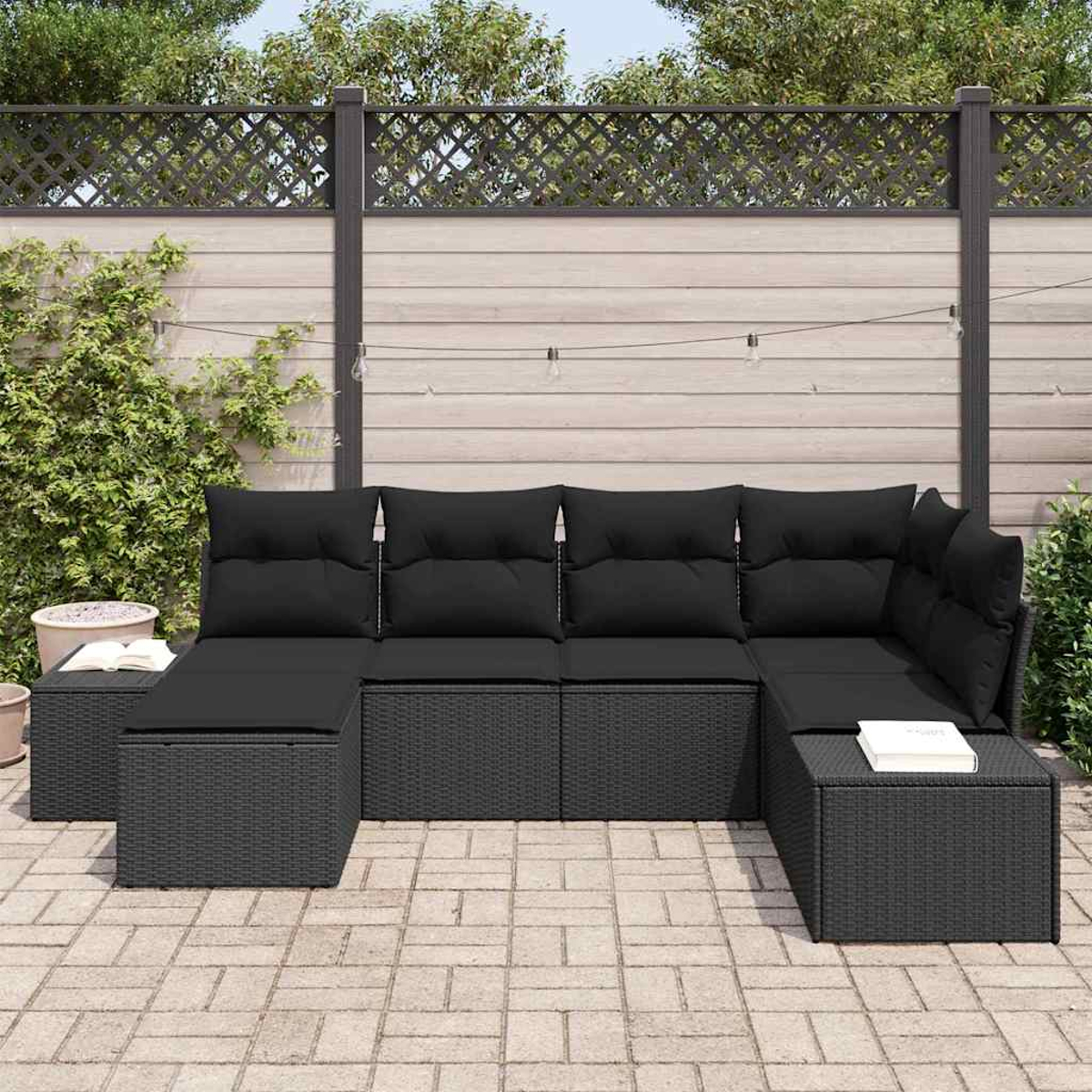 Divano angolare in vimini nero con cuscini neri su un patio pavimentato. Recinzione in legno e verde sullo sfondo.