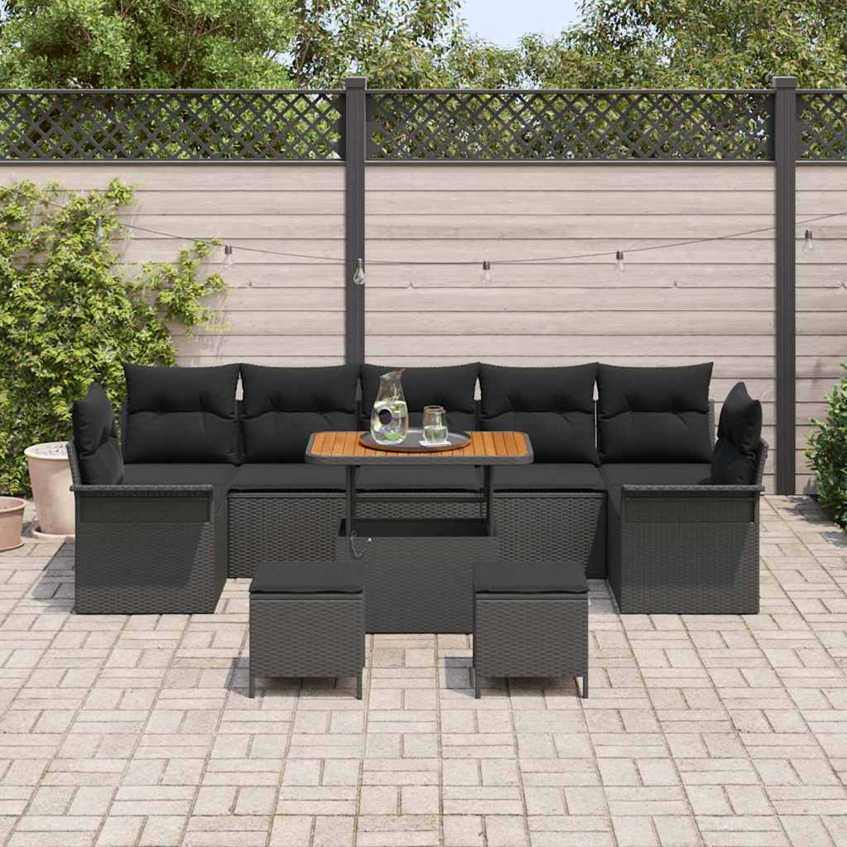 Juego de asientos al aire libre con cojines negros, una mesa de madera y dos otomanas. El conjunto está sobre un patio de ladrillos con una valla de madera.