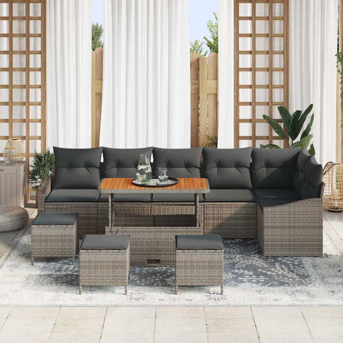 Outdoor-Ecksofa mit grauen Kissen, einem Holztisch und Hockern auf einem Teppich. Weiße Vorhänge und ein Holzzaun im Hintergrund.