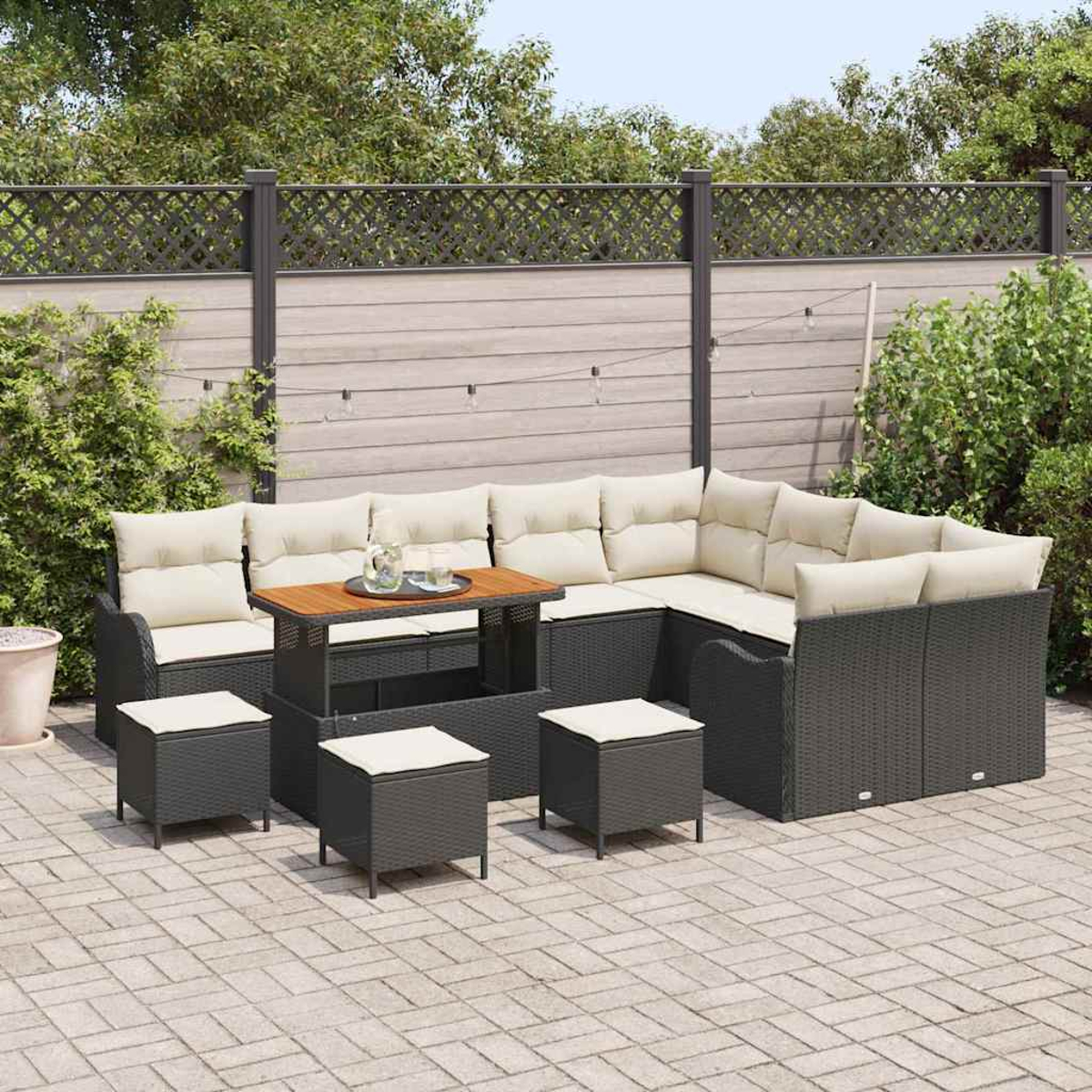 Garten-Terrassenset mit großem Ecksofa, Ottomanen und Tisch auf gepflasterter Terrasse. Im Hintergrund Zaun und Grün.