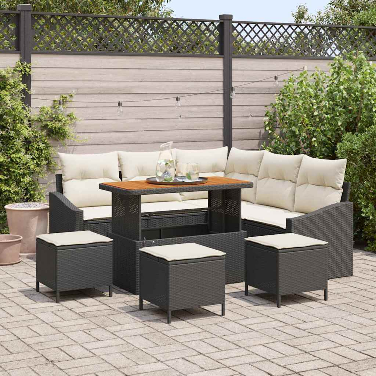 Gartenmöbel-Set aus Korbgeflecht mit Tisch, Sofa und Hockern auf einer Ziegelterrasse, mit Holzzaun und Grün im Hintergrund.