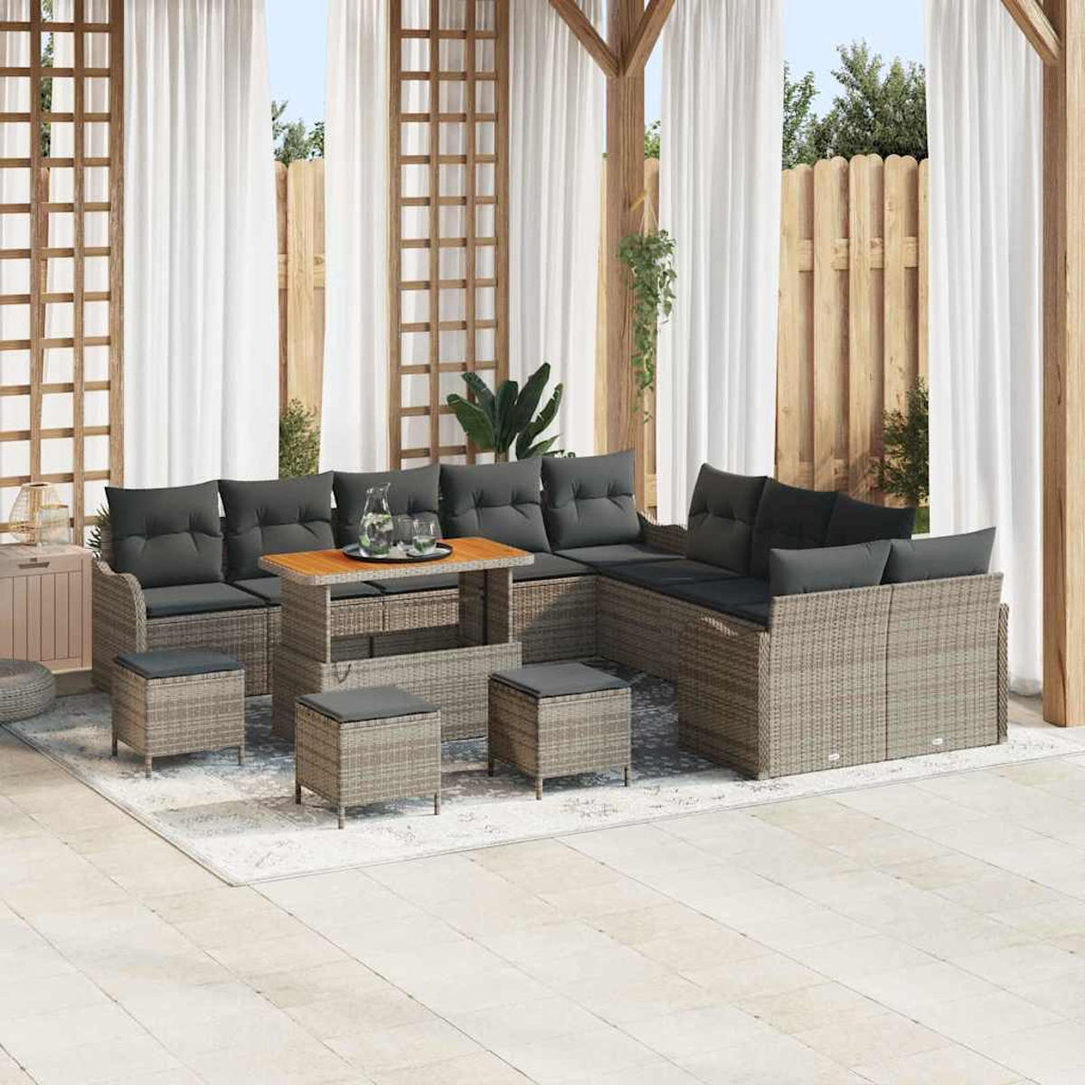 Ein graues Outdoor-Ecksofa aus Korbgeflecht mit schwarzen Kissen, ein Holztisch und Hocker stehen auf einem Teppich unter einer Pergola.