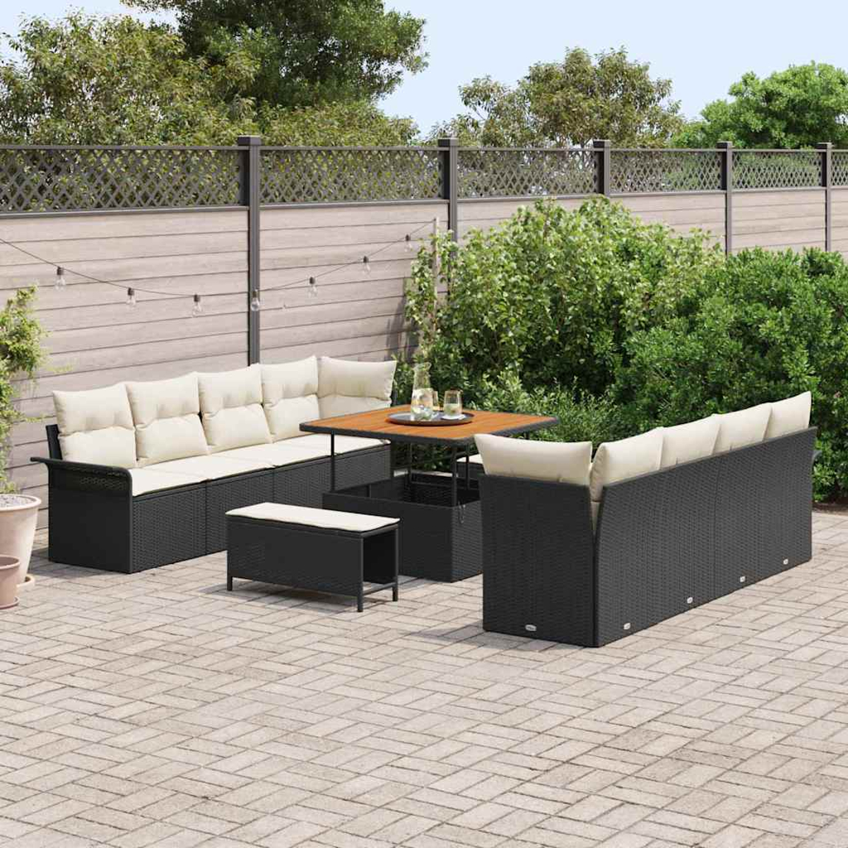 Set da patio esterno con divani e panca in vimini neri, cuscini beige, tavolo in legno e patio in mattoni, il tutto contro una recinzione e vegetazione.