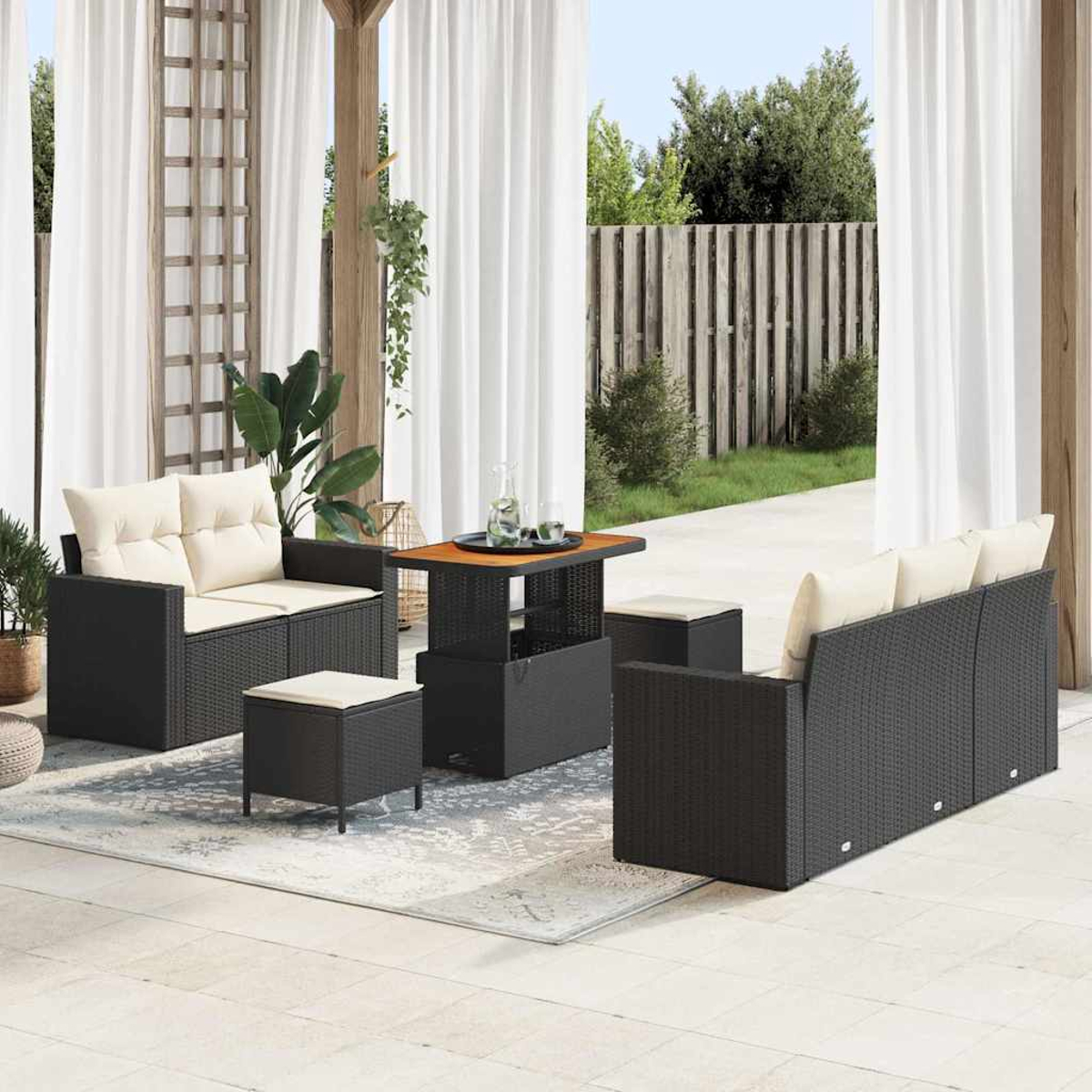 Set da patio esterno con mobili in vimini neri, cuscini beige e un tavolo di legno. Tende bianche e una staccionata in legno sullo sfondo.