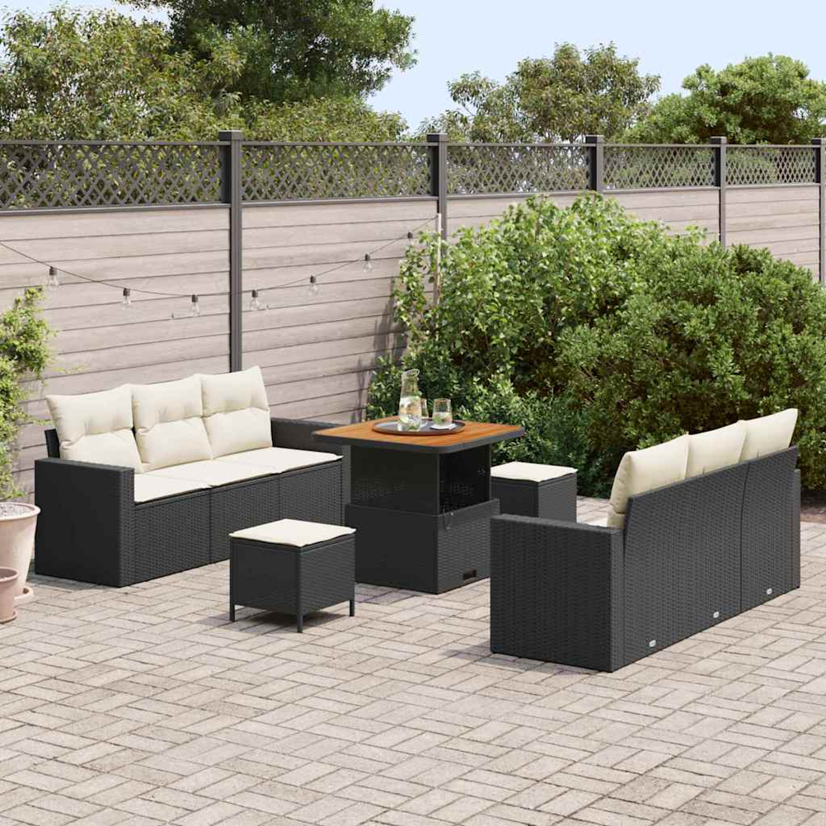 Conjunto de patio exterior: muebles de mimbre negro con cojines crema, una mesa con bebidas, sobre un patio de piedra, valla al fondo.