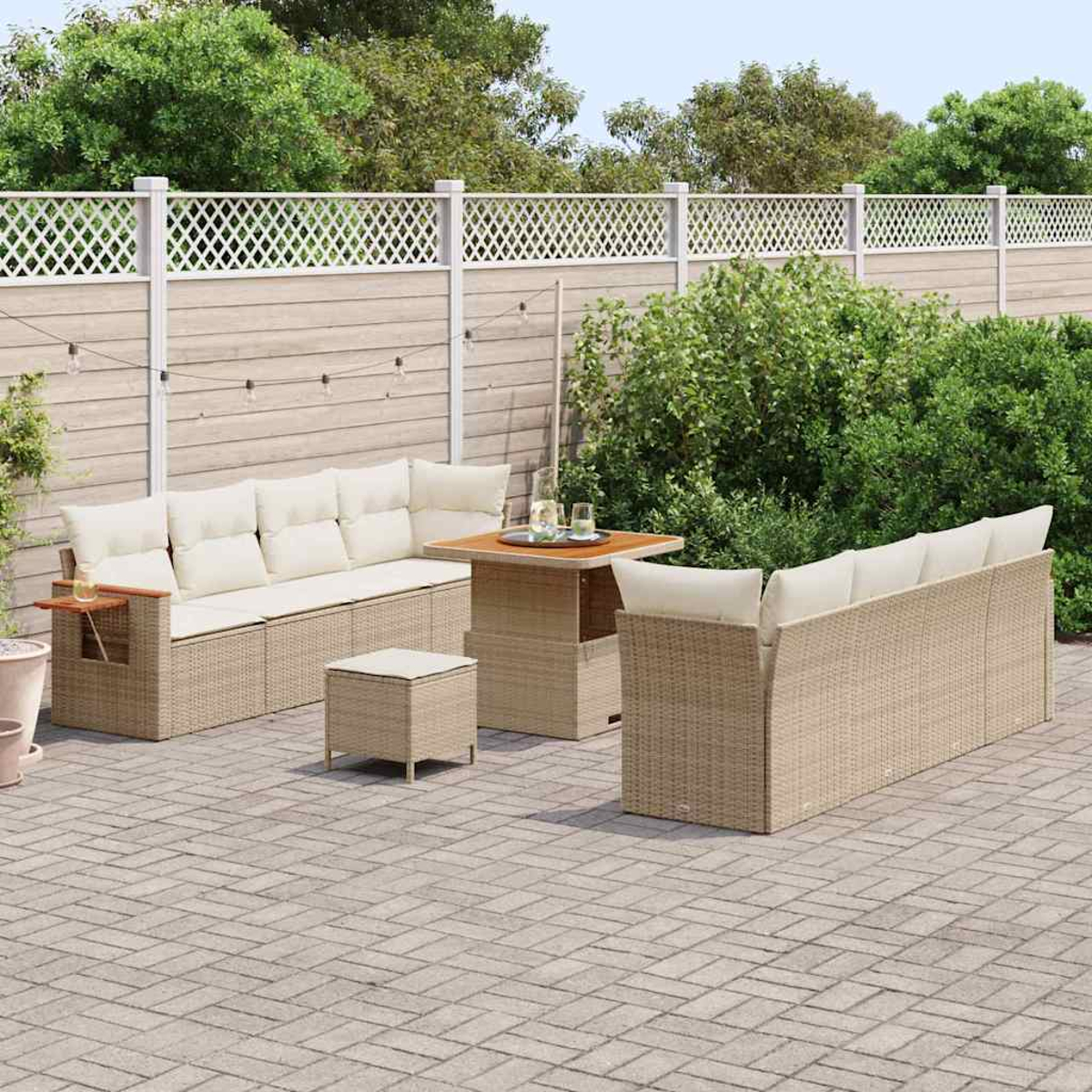 Set da patio esterno con divani in vimini beige, un tavolo e un piccolo pouf, su un patio in mattoni. Piante verdi e una recinzione bianca.