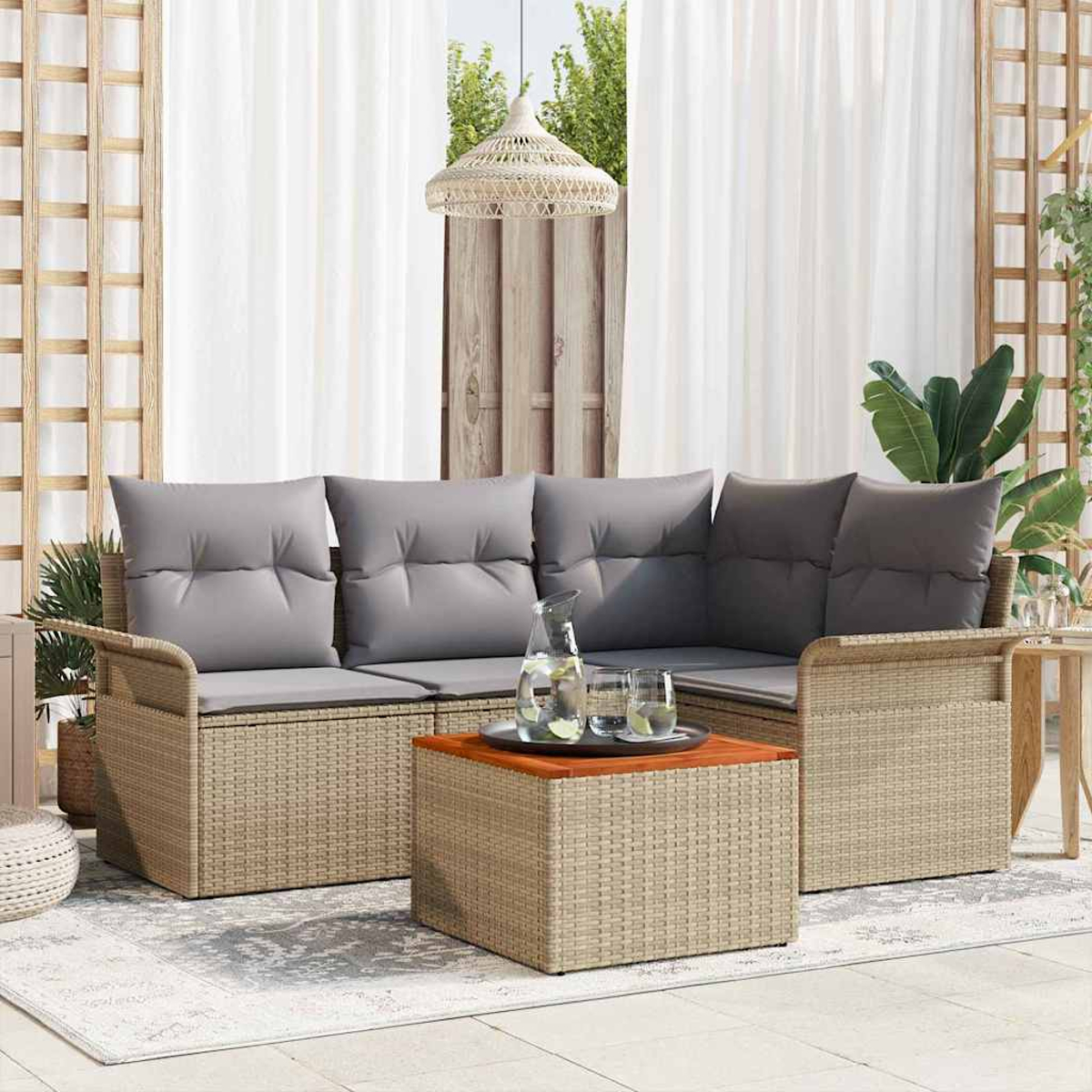 Outdoor-Sitzbereich mit beigem Websofa, grauen Kissen, Holztisch und Wasserkrug. Vorhänge und Grün im Hintergrund.