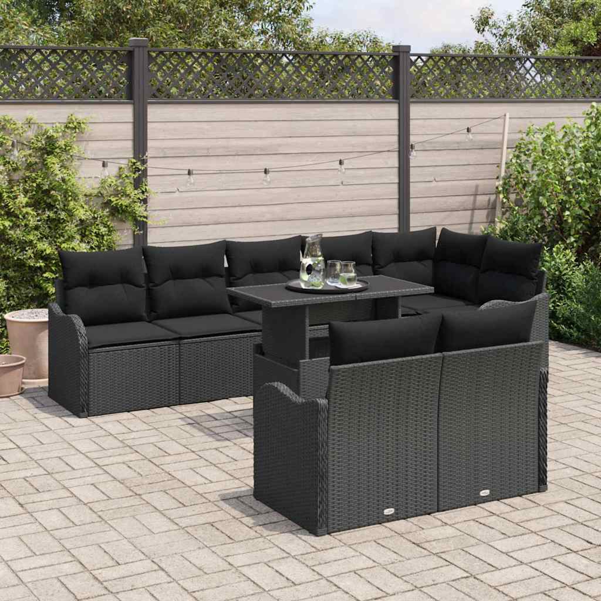 Szene im Freien mit schwarzen Korbmöbeln, Tisch mit Getränken, Zaun und Pflanzen. Gepflasterte Terrasse mit Ziegelmuster.