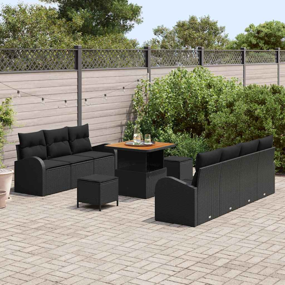 Garten-Terrassenset mit schwarzen Korbmöbeln. Enthält Sofa, Loveseat, Tisch und Ottomanen auf einer gepflasterten Terrasse, mit Grün und Zaun.