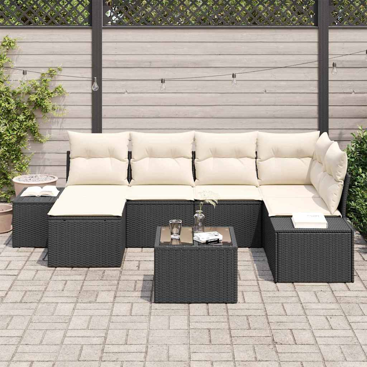 Outdoor-Ecksofa mit beigen Kissen und schwarzem Korbgewebe. Davor steht ein kleiner Tisch mit Glasplatte. Im Hintergrund ein Holzzaun.