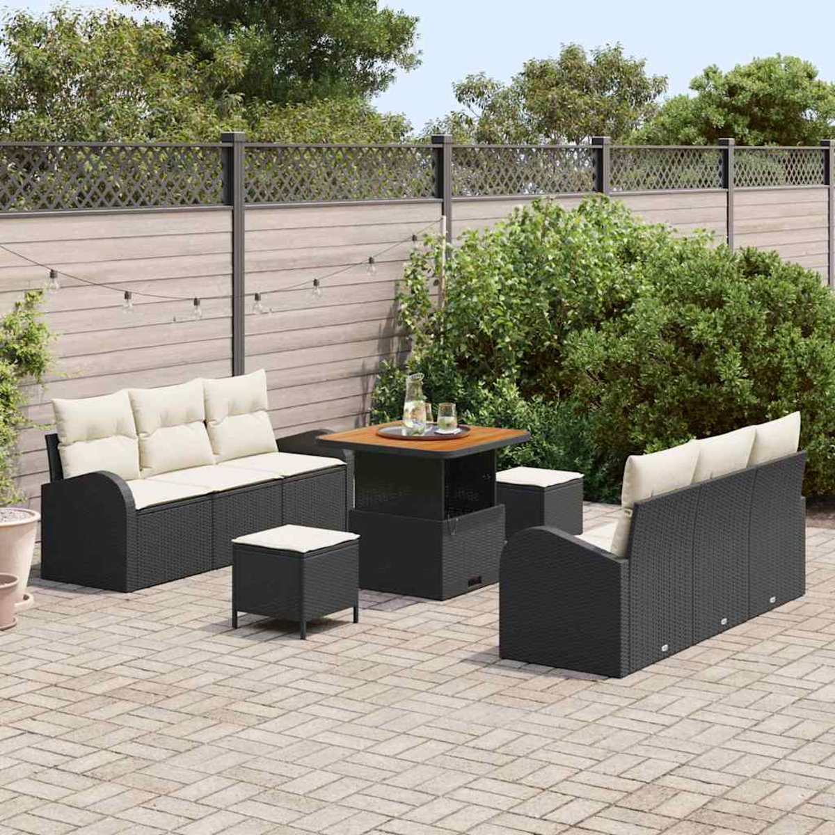 Terrassengarnitur: Schwarze Korbmöbel mit cremefarbenen Kissen, Tisch und zwei Ottomanen. Holzzaun und Grün im Hintergrund.
