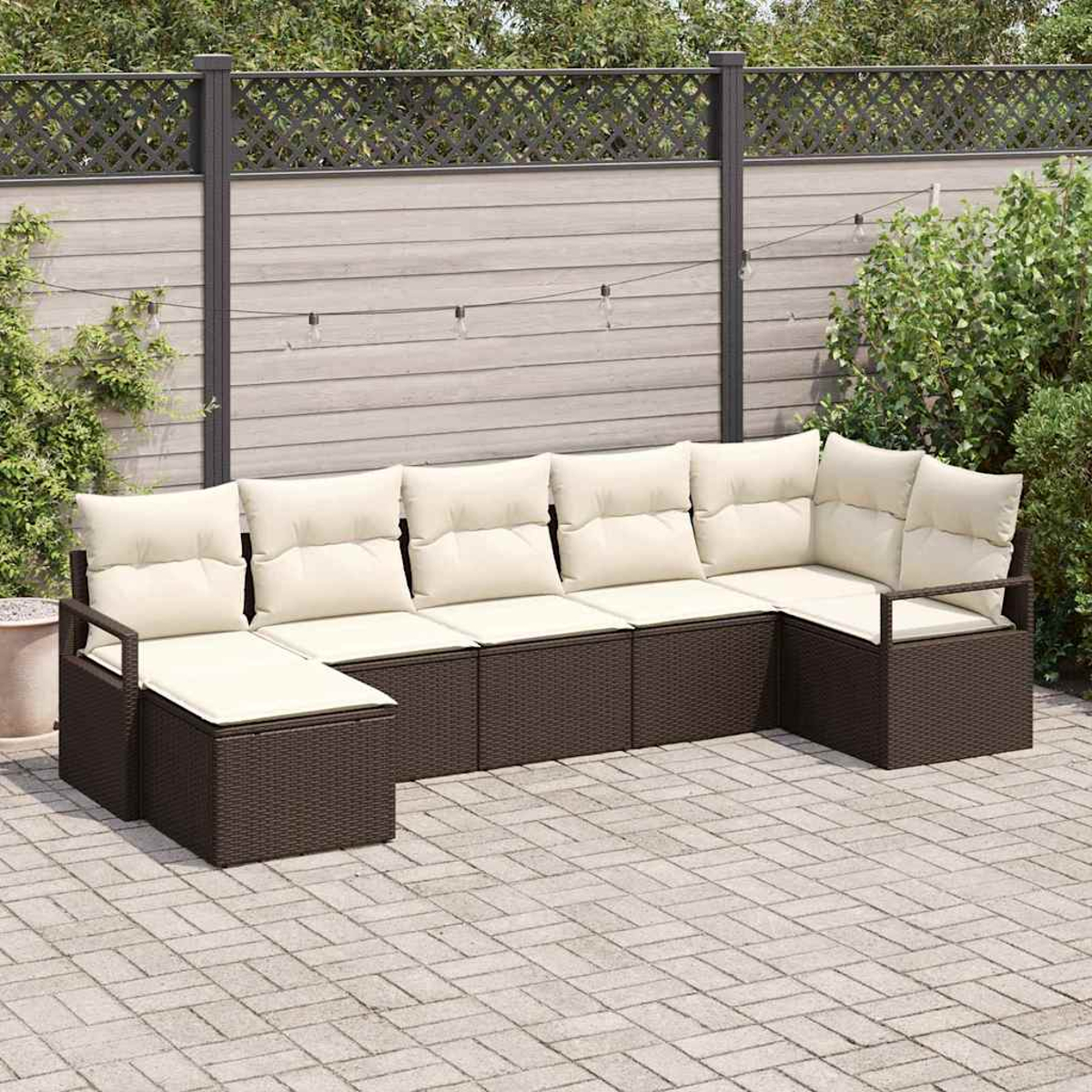 Braunes Rattan-Outdoor-Ecksofa mit beigen Kissen auf einer gepflasterten Terrasse. Ein Zaun und Grün bilden den Hintergrund.