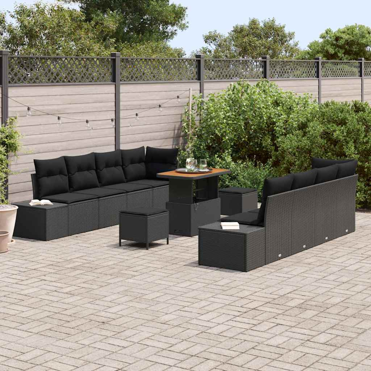 Terrassengarnitur mit schwarzen Kissen, Tisch und Sitzgelegenheiten. Ein Holzzaun und grüne Büsche im Hintergrund.