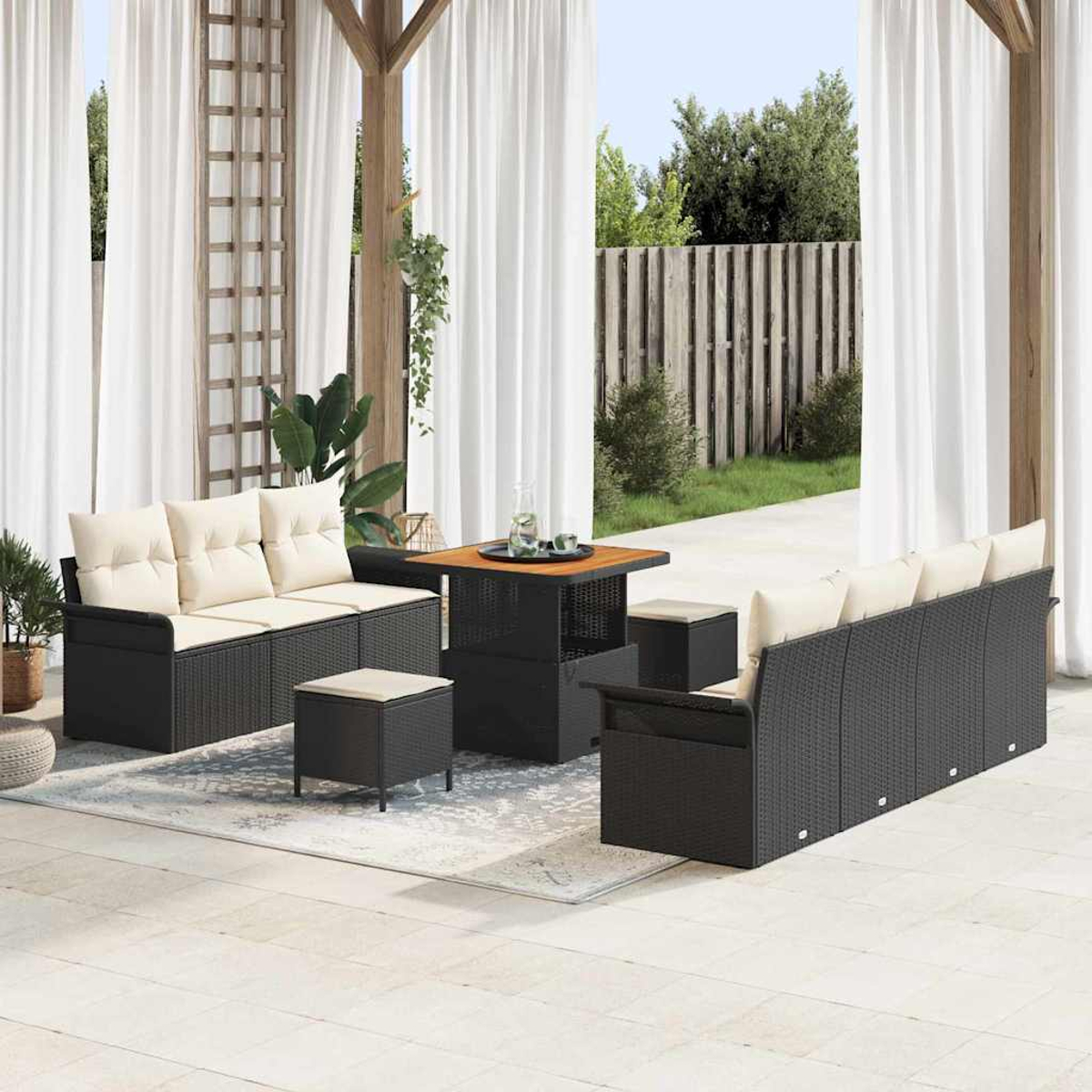 Juego de patio al aire libre con muebles de mimbre negro, cojines crema y una mesa de madera. Cortinas blancas y vegetación al fondo.