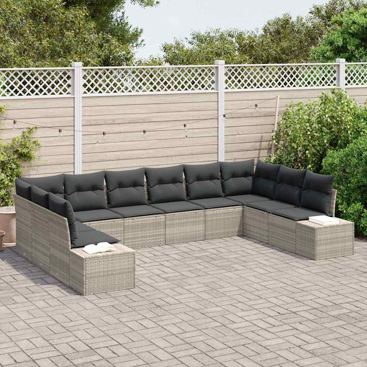 Ein graues Rattansofa mit dunklen Kissen steht auf einer Ziegelterrasse. Ein weißer Zaun und grüne Büsche bilden den Hintergrund.