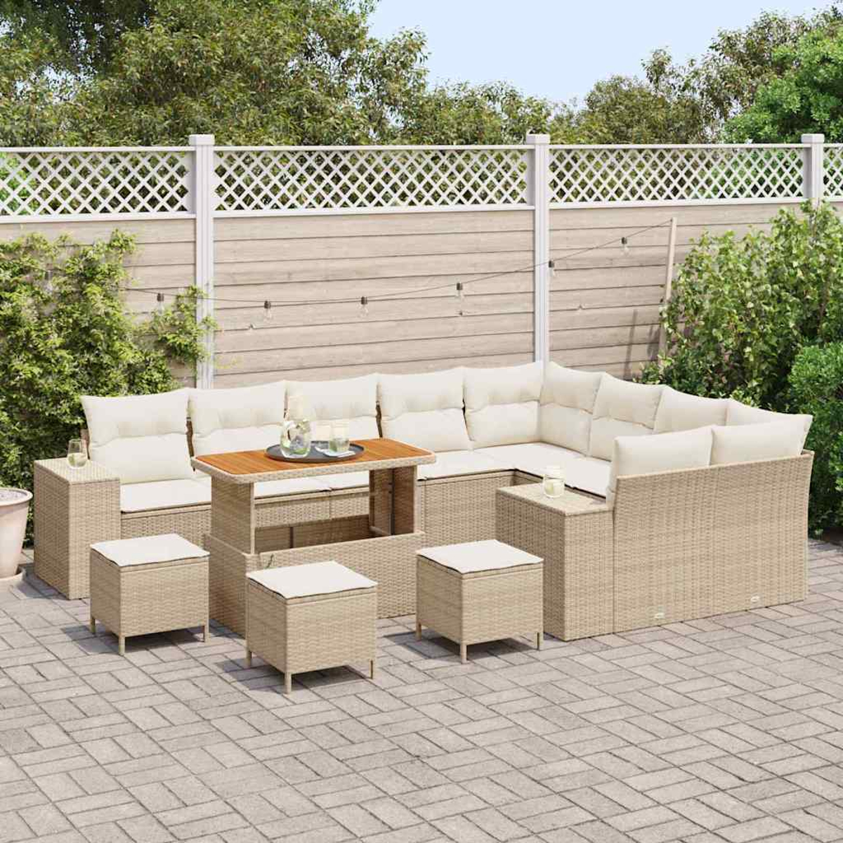 Gartenmöbel-Set aus Korbgeflecht mit beigen Kissen, einem Holztisch und passenden Hockern auf einer gepflasterten Terrasse, mit Zaun und Grün im Hintergrund.
