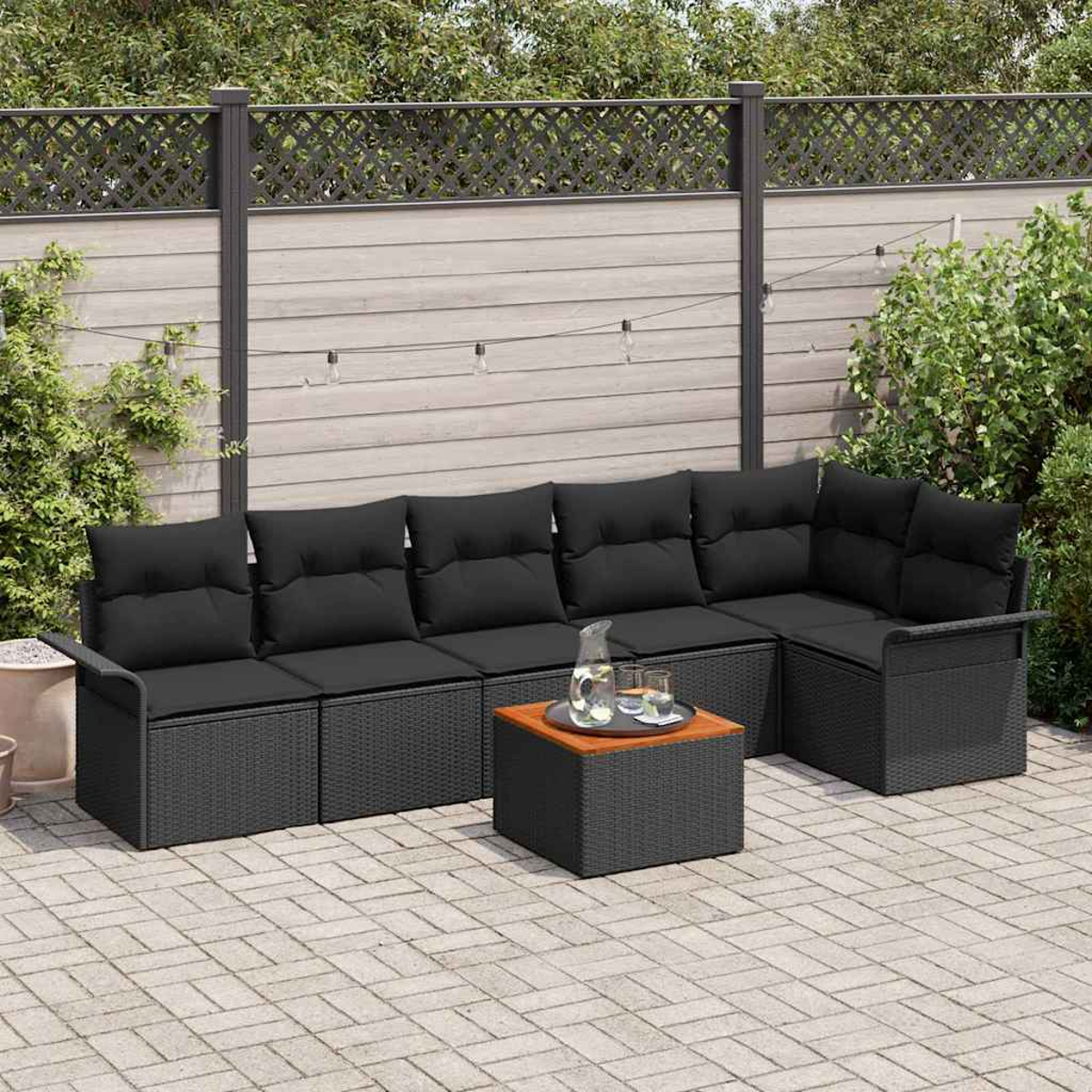 Un sofá seccional negro con cojines, una mesita con bebidas y una valla al fondo. Patio pavimentado.