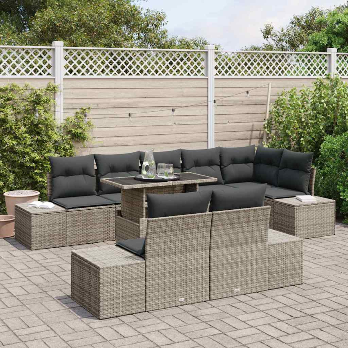 Gartenmöbel-Set aus Rattan mit dunkelgrauen Kissen auf einer Terrasse. Ein Tisch mit Gläsern und einer Kanne steht in der Mitte. Ein weißer Zaun im Hintergrund.