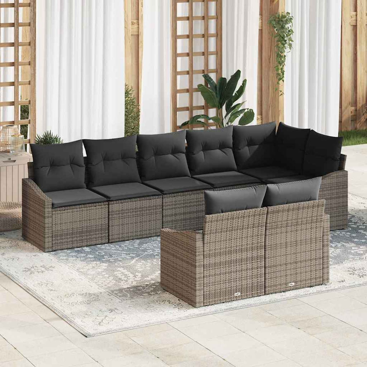 Outdoor-Ecksofa mit grauen Kissen. Korbgewebe-Rahmen, auf einem gemusterten Teppich. Weiße Vorhänge, Holzgitter und Pflanzen im Hintergrund.