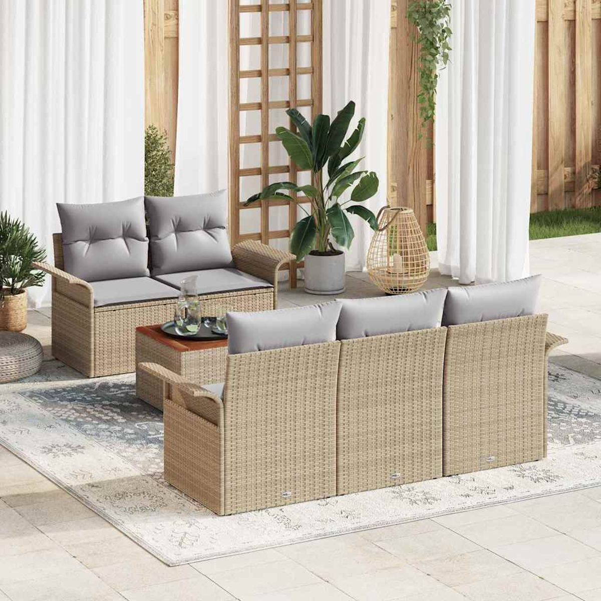 Gartenmöbel-Set aus Rattan mit grauen Kissen, Tisch, Pflanzen und Teppich auf einer Terrasse. Weiße Vorhänge und Holzgitter im Hintergrund.