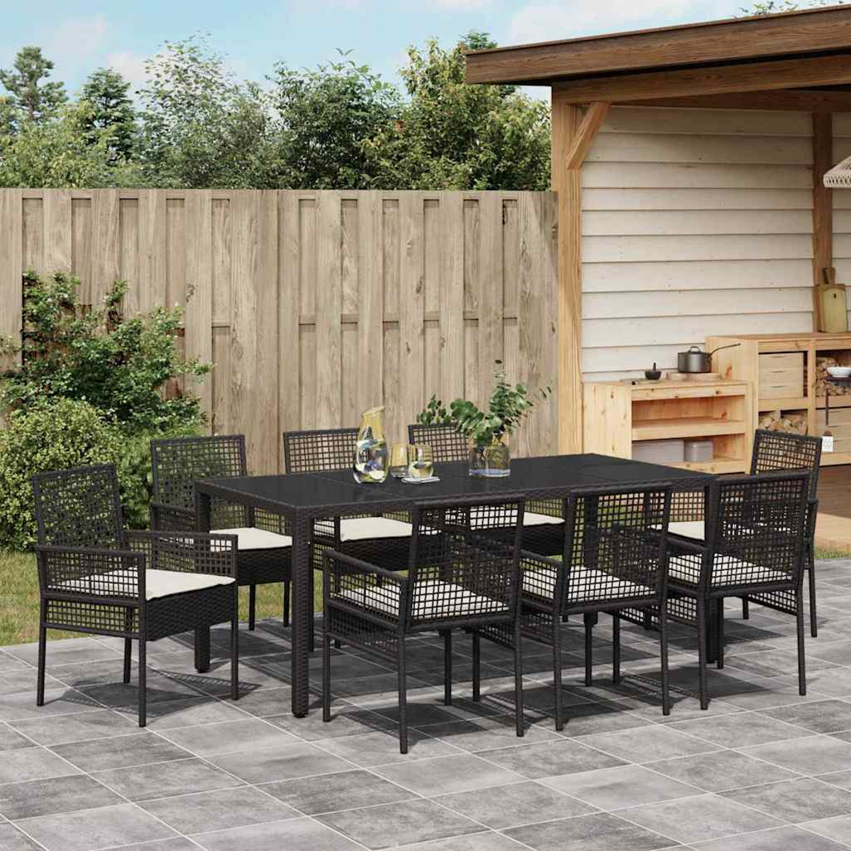 Ein dunkler Tisch mit Stühlen steht auf einer Terrasse. Ein Holzzaun und eine kleine Holzkonstruktion sind im Hintergrund.