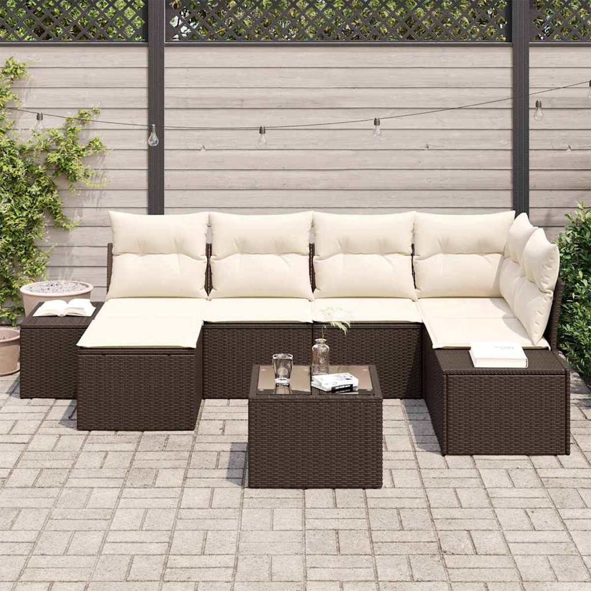 Divano angolare esterno in vimini marrone con cuscini color crema, tavolo di vetro, libri e bevande. Recinzione in legno e piante sullo sfondo.