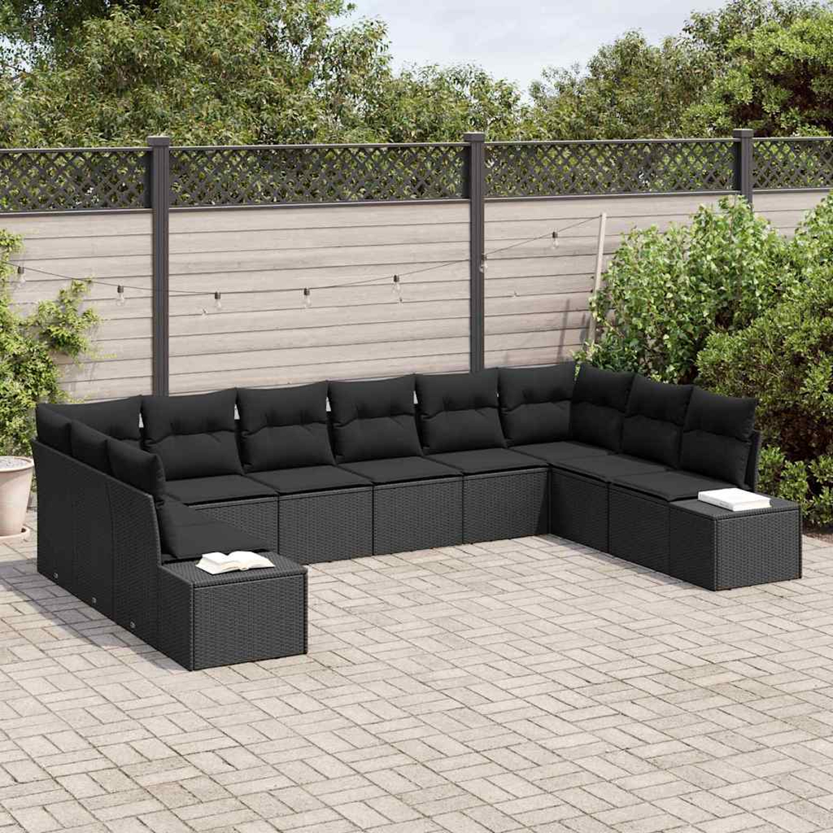 U-förmiges schwarzes Rattan-Sofa mit Kissen auf einer Terrasse, umgeben von Grün und einem Holzzaun.