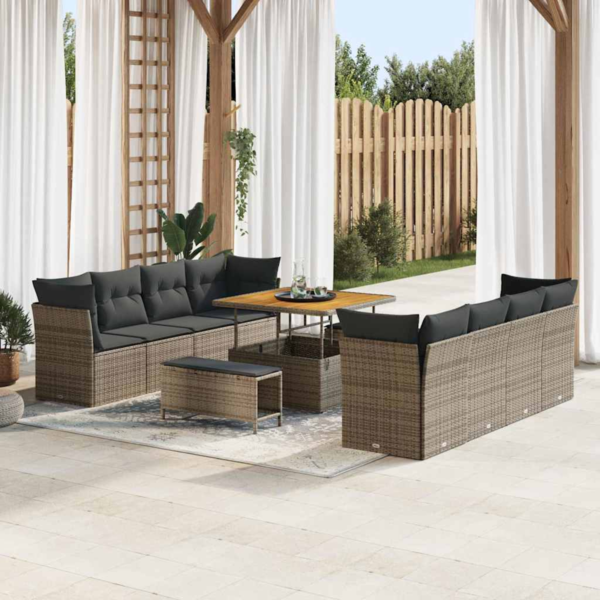 Terrassenset im Freien mit braunen Korbs Sofas, grauen Kissen, Holztisch und weißen Vorhängen. Im Hintergrund ein Holzzaun und Bäume.