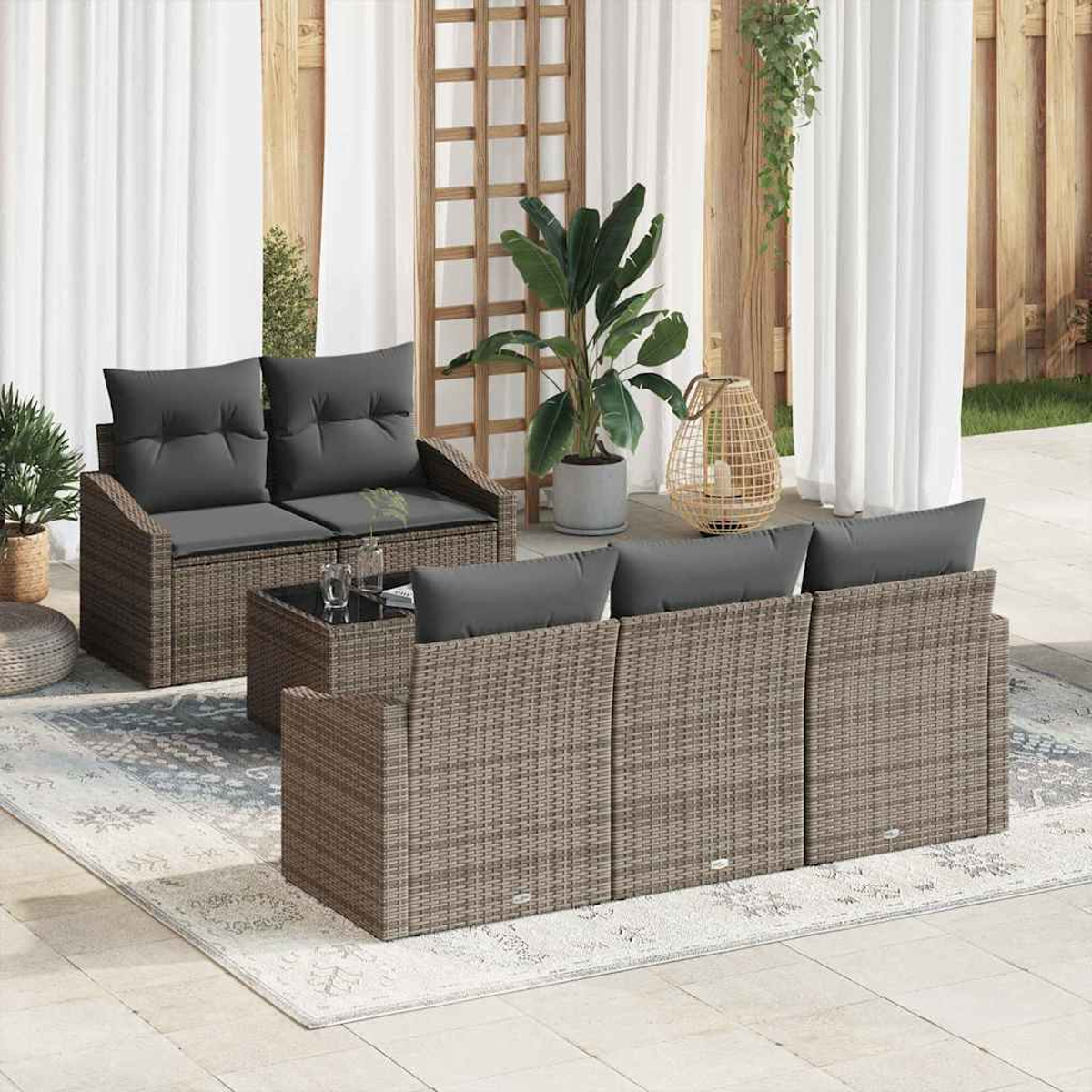 Gartenmöbel-Set aus Korbgeflecht mit grauen Kissen auf einem Teppich. Pflanzen und ein Gitter-Hintergrund. Neutrale Töne, helles Sonnenlicht.