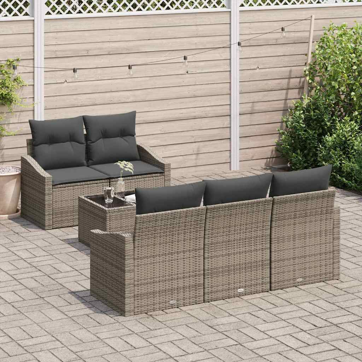 Gartenmöbel-Set mit grauen Kissen, einem kleinen Tisch und einem Holzzaun. Gepflasterte Terrasse mit Pflanzen und Lichterketten.