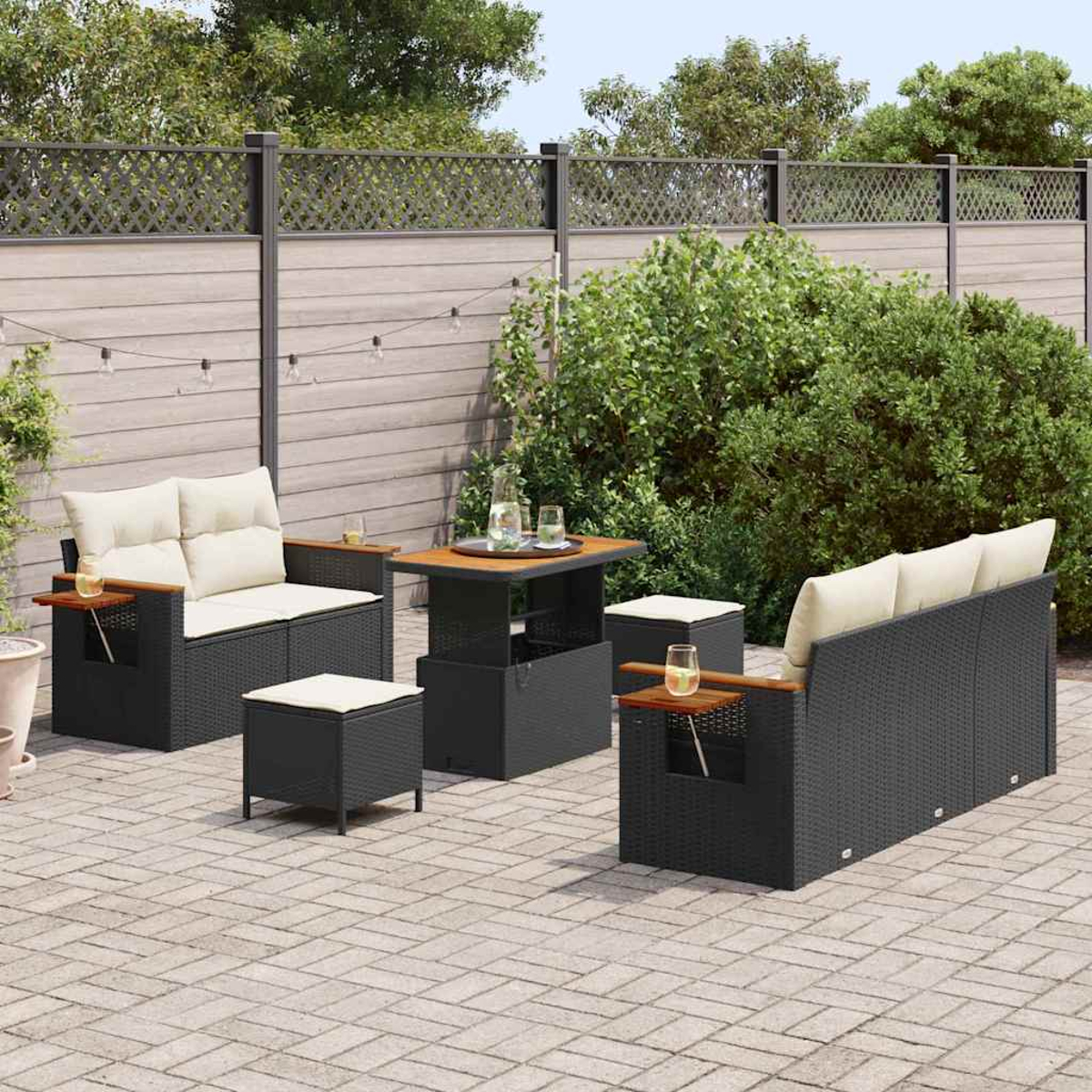 Set da patio all'aperto con mobili in vimini neri, cuscini beige e un tavolo in legno. Il set è su un patio in mattoni con verde e una recinzione.