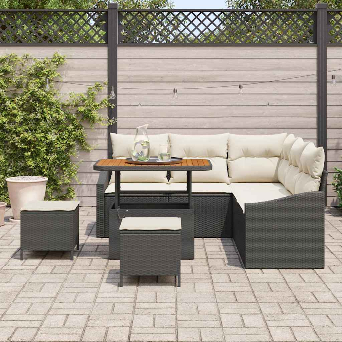 Outdoor-Sitzgruppe mit Ecksofa, Tisch und zwei Hockern auf einer Ziegelsteinterrasse. Im Hintergrund ein Holzzaun und Grün.