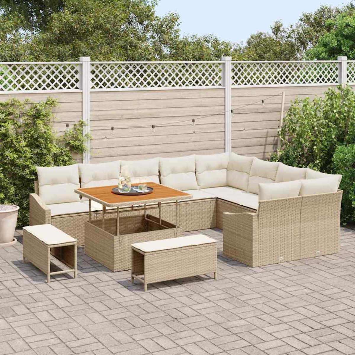 Divano angolare in rattan da esterno con cuscini, un tavolo e due panche su un patio. Una recinzione e vegetazione sullo sfondo.
