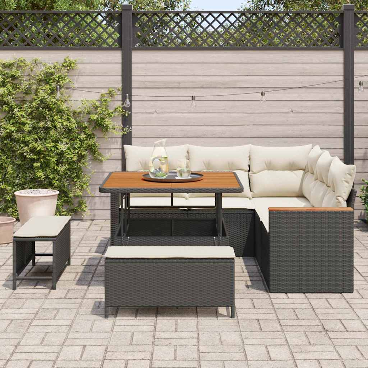 Juego de patio al aire libre con mesa, sofá seccional y banco. Mesa y estructura de sofá de madera. Cojines de color claro. Plantas verdes y valla al fondo.