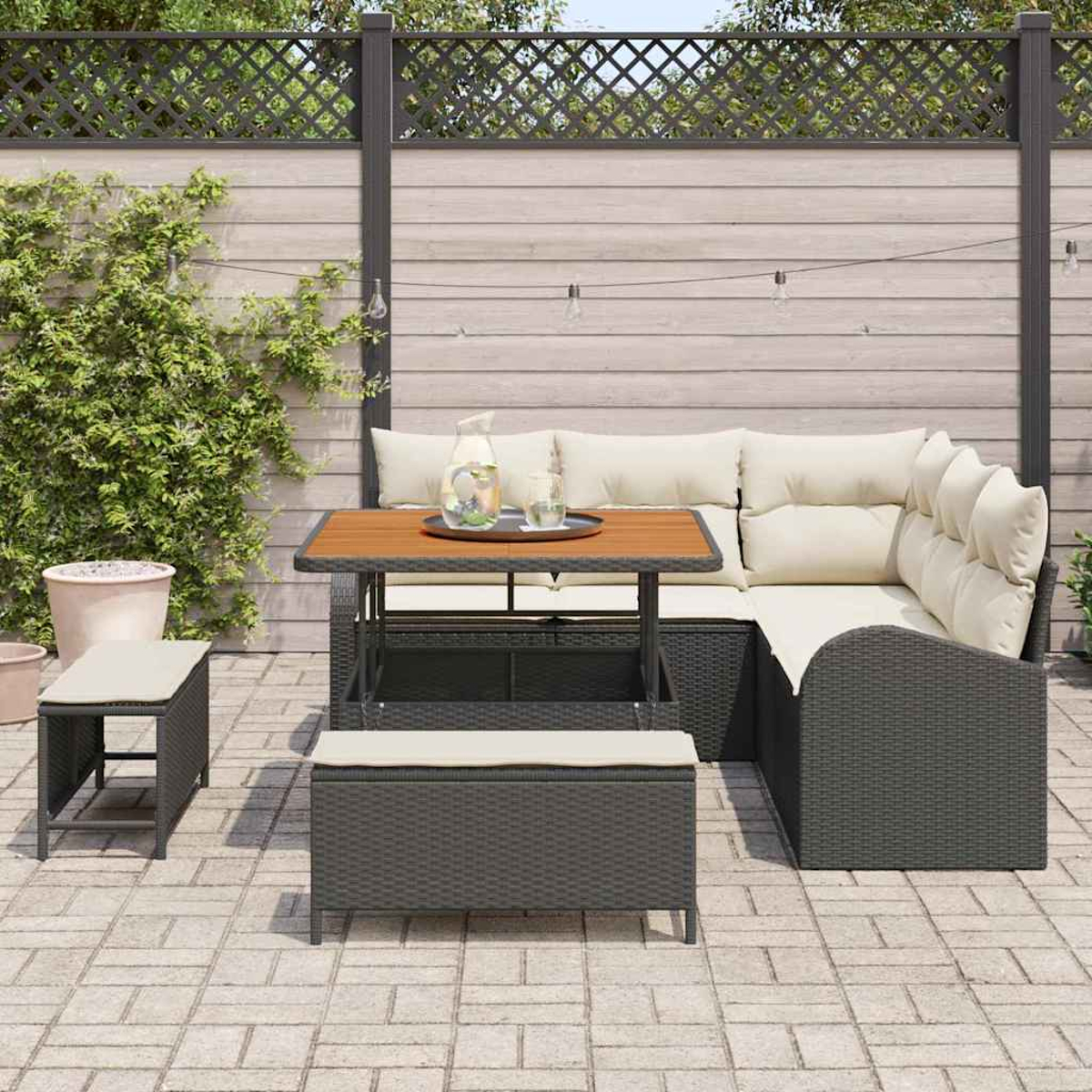 Juego de patio al aire libre con sofá de mimbre, mesa de madera y bancos acolchados. Una jarra y vasos están sobre la mesa. Una valla de madera al fondo.