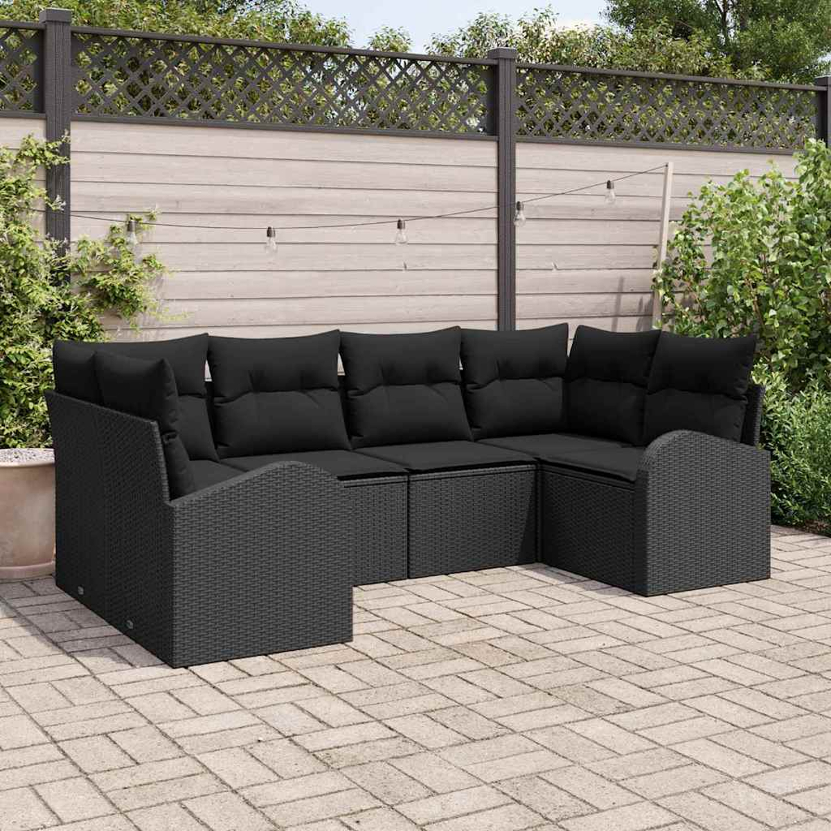 Ein schwarzes Korbsessel-Ecksofa mit schwarzen Kissen steht auf einer Terrasse. Dahinter befinden sich ein Zaun und Grün, mit Lichterketten.