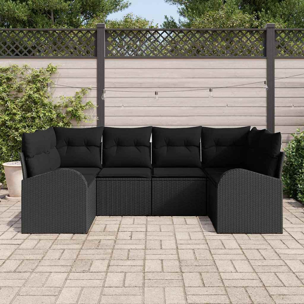 Schwarzes Outdoor-Sofa aus Korbgeflecht auf einer gepflasterten Terrasse. Ein Holzzaun mit Kletterpflanzen und Lichterketten im Hintergrund.