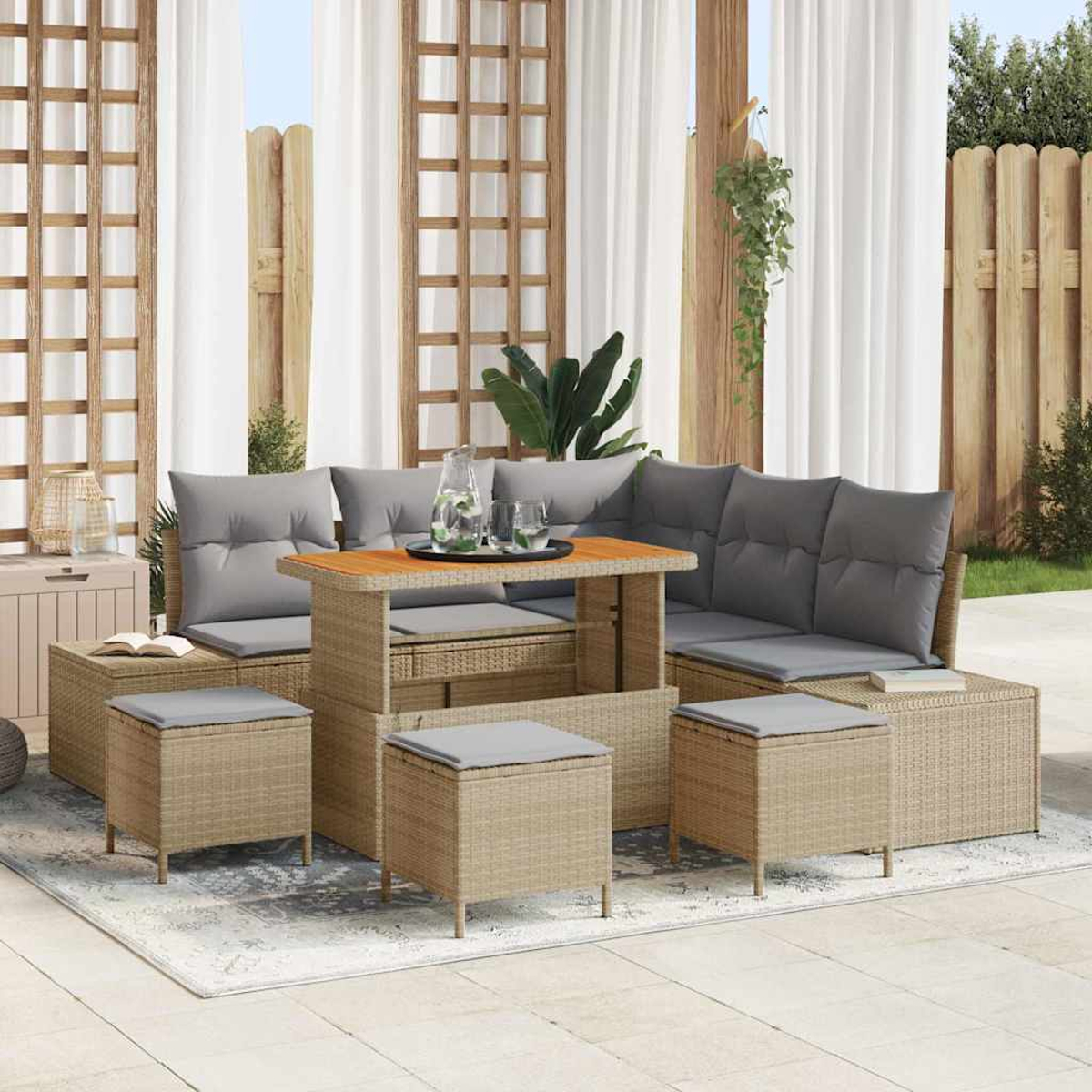 Gartenlounge-Set mit Tisch, Sofas und Hockern. Graue Kissen auf Rattanmöbeln. Weiße Vorhänge und Holzzaun im Hintergrund.