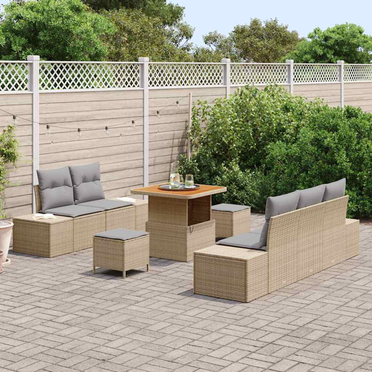 Gartenmöbel-Set aus Korbgeflecht mit grauen Kissen, einem quadratischen Tisch und Hockern auf einer Backstein-Terrasse, umgeben von Grün und einem Zaun.