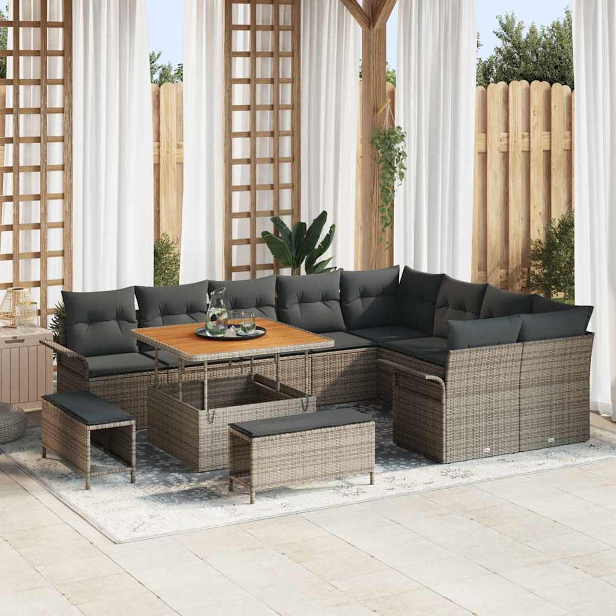 Set da patio con cuscini grigi, tavolo in legno e panche su tappeto, tende e staccionata in legno sullo sfondo.