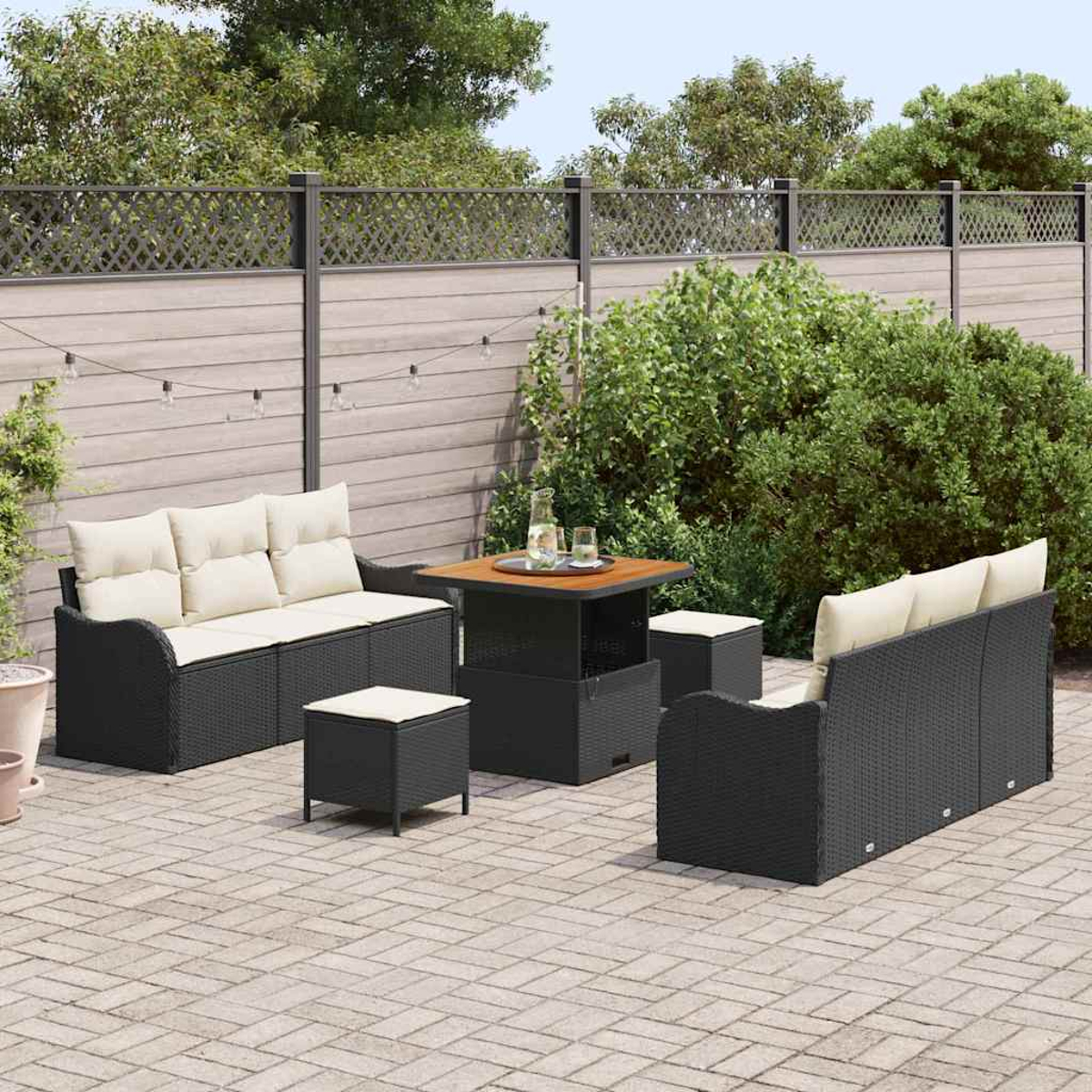 Gartenmöbel-Set. Schwarze Korbsessel, Hocker und Tisch mit cremefarbenen Kissen auf einer Ziegelterrasse, Zaun im Hintergrund.