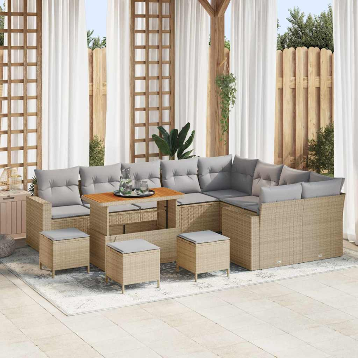 Outdoor-Sitzgruppe mit grauen Kissen, einem Holztisch und Hockern auf einem gemusterten Teppich. Durchscheinende Vorhänge und ein Holzzaun im Hintergrund.