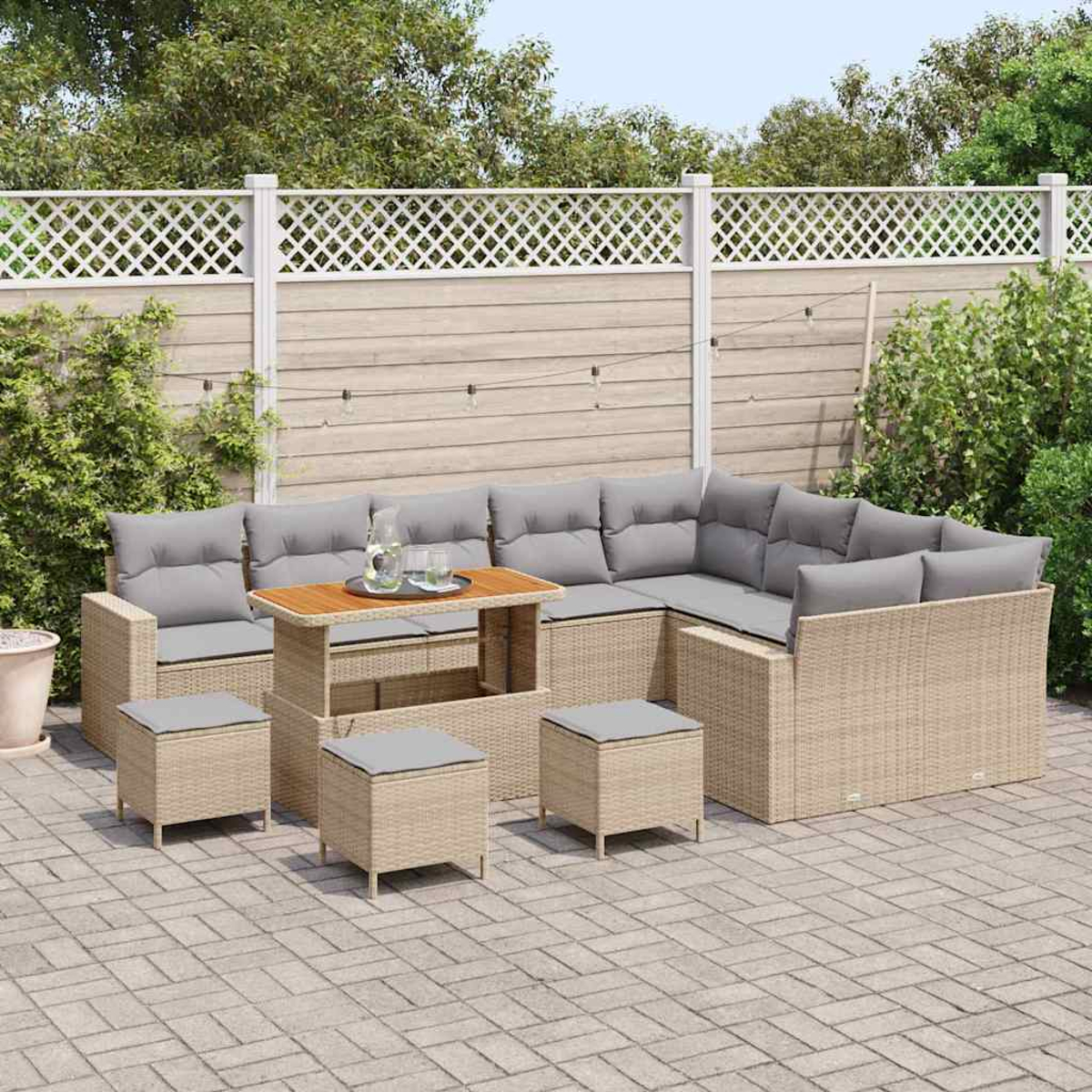 Garten-Terrassenset mit beigen Korbmöbeln, grauen Kissen und einem Holztisch. Auf einer Steinterrasse mit weißem Zaun und Grün.